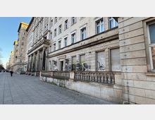 Lange, mehrstöckige Sandsteinfassade eines historischen Gebäudekomplexes entlang eines breiten Gehwegs, mit hohen Säulen am Eingangsbereich. Im Erdgeschoss sind mehrere Fenster mit Rollläden sowie Graffiti zu sehen, davor eine niedrige Balustrade aus Stein.