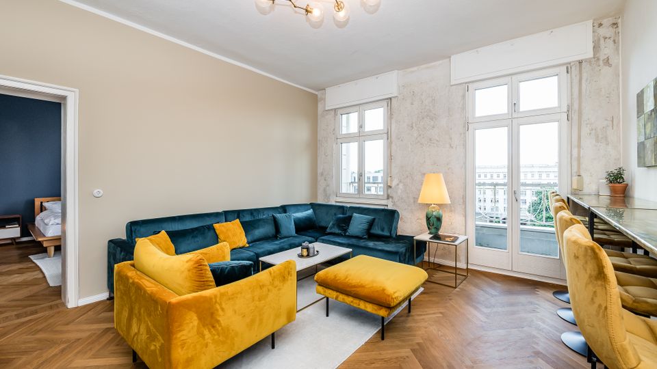 Modernes Wohnzimmer mit Fischgrätparkett, großem L-förmigem Sofa und gelbem Sessel samt Ottomane um einen Couchtisch. Hohe Fenster und eine Balkontür lassen viel Tageslicht herein; rechts steht eine schmale Theke mit Barhockern unter einer Deckenleuchte.