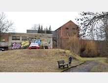 Blick auf ein niedriges, graues Gebäude mit Graffiti und Treppenaufgang, daneben ein höherer Backsteinbau. Davor liegt eine grasige Böschung mit Parkbank und einem Weg, umgeben von kahlen Bäumen und Sträuchern.