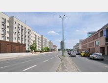 Straßenszene in einer Stadt mit mehrspuriger Fahrbahn, flankiert von modernen, mehrstöckigen Wohn- und Geschäftsgebäuden. Rechts parkende und fahrende Autos, links Gehweg mit vereinzelten Bäumen vor einem Wohngebäude.