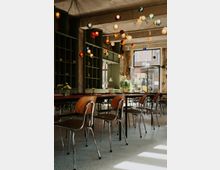 Ein modernes Café mit industriellem Design, erkennbar an den sichtbar belassenen Betonwänden und Decken. Bunte hängende Glühlampen schmücken die Decke, während Holztische und -stühle die Einrichtung dominieren; große Fenster lassen viel Tageslicht herein.