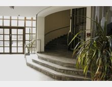 Eingangsbereich eines Gebäudes mit breiten Marmorstufen, großen Fenstern mit Holzrahmen und einer geschwungenen Treppe im Hintergrund. Pflanzendekoration, darunter ein Kaktus und eine große Grünpflanze, ist am Rand zu sehen.