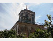 Historisches Gebäude mit oktogonalem Turm, verziert mit gotischen Fenstern und Balustraden. Im Vordergrund sind Bäume und Sträucher, der Himmel ist blau mit leichten Wolken.