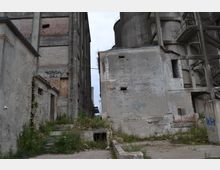 Industriegelände mit verfallenen Gebäuden aus Beton und Ziegelstein, teils mit Graffiti bedeckt; überwucherte Vegetation zwischen den Bauten und beschädigte Treppenstufen im Vordergrund.