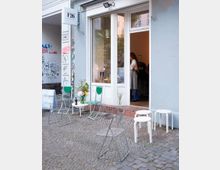 Ein kleiner Laden mit einer großen Glastür und weißen Rahmen an einer hellblauen Fassade. Vor dem Eingang stehen verschiedene einfache Stühle und Hocker auf einem Pflastersteinboden, daneben befindet sich etwas Graffiti an der Wand.