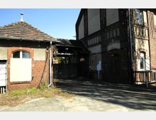 Außenansicht eines verfallenen Backsteinbaus mit teilweise verputzten Wänden. Links ein kleines Gebäude mit Ziegeldach und zugemauertem Fenster, rechts ein größeres Gebäude mit hohen Fenstern und sichtbarem Verfall, umgeben von einem gepflasterten Hof.
