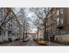 Eine ruhige Wohnstraße mit mehrstöckigen Altbauten auf beiden Seiten. Die Bäume ohne Laub säumen die Straße, auf der parkende Autos stehen; am Ende der Straße ist ein weiteres Gebäude mit rotem Dach zu sehen.