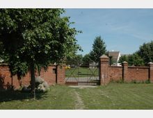 Eine Ziegelmauer mit einem eisernen Doppeltor in der Mitte, das zu einer Wiese mit Bäumen und einem dahinterliegenden Gebäude führt. Im Vordergrund befindet sich ein Baum und eine Rasenfläche.