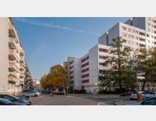 Straßenszene in einem Wohngebiet mit mehrstöckigen Wohngebäuden auf beiden Seiten der Straße, teils mit Balkonen. Der Vordergrund zeigt parkende Autos, einen Fußgängerüberweg und Straßenschilder, während Bäume und Herbstlaub das Bild ergänzen.