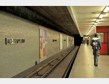 U-Bahn-Station "Alt-Tempelhof" mit einem leeren Gleis und einem Bahnsteig. An den Wänden befinden sich Kacheln, Werbung und Stationsschilder; die Decke ist aus Beton und mit Lichtern beleuchtet, während Personen auf dem Bahnsteig zu sehen sind.