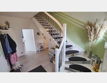 Treppenhaus in einem modern eingerichteten Wohnraum mit weißer Wendeltreppe und metallenen Geländern. Die Wände sind grün mit weißen Streifen dekoriert, und der Raum enthält Dekorationen wie Pampasgras in einer Vase, Laternen und eine Garderobe.