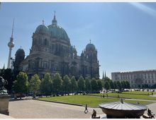 Berliner Dom mit seinen markanten grünen Kuppeln und detaillierter Fassade, umgeben von gepflegten Rasenflächen und Bäumen, im Hintergrund der Berliner Fernsehturm und moderne Gebäude. Im Vordergrund ein Brunnen mit rundem Dach.