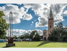 Eine historische Kirche mit einem hohen Glockenturm und Spitzdach steht auf einer offenen, grasbewachsenen Fläche, flankiert von Bäumen. Links im Vordergrund ist eine schlanke, moderne Metallskulptur, während im Hintergrund geparkte Autos und teils bewölkter Himmel zu sehen sind.