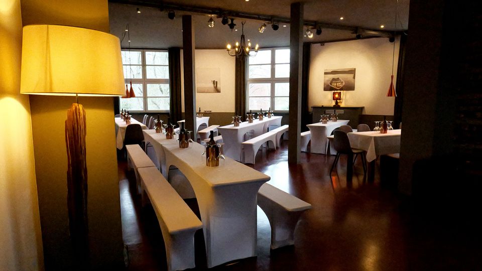 Innenraum eines Restaurants mit langen, weiß gedeckten Tafeln und Bänken sowie weiteren Tischen mit Stühlen auf dunklem Holzboden. Große Sprossenfenster, schwarze Stützsäulen, eine Kronleuchte und Pendelleuchten prägen den Raum.