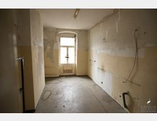 Ein leerer, renovierungsbedürftiger Raum mit abblätternden Wänden in beige und weiß, sichtbaren Rohrleitungen und freiliegenden Kabeln. Im Hintergrund befindet sich ein einzelnes, schmutziges Fenster mit Tageslichteinfall.