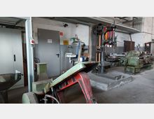 Eine Werkstatt mit alten Maschinen und Werkzeugen, einschließlich einer Bohrmaschine und einer Drehbank. Im Hintergrund sind ein Waschbecken, eine graue Tür und Regale mit Werkzeugen zu sehen.