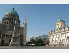 Historischer Platz mit der St. Nikolaikirche links, erkennbar an ihrer großen grünen Kuppel und Säulenfassade, sowie dem Barockbau des Alten Rathauses rechts mit goldener Figur auf der Kuppel. In der Mitte steht ein Obelisk und im Hintergrund sind Bäume und moderne Gebäude zu sehen.
