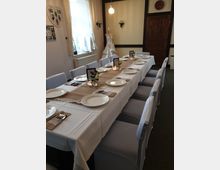 Ein festlich gedeckter Tisch in einem hellen Raum mit weißen Tischdecken, Geschirr und dekorativen Elementen wie Kerzen und Blumen. Im Hintergrund sind eine Schaufensterpuppe mit einem weißen Kleid, eine Holztür sowie ein Fenster mit Vorhängen zu sehen.