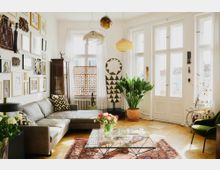 Helles Wohnzimmer mit L-förmigem grauem Sofa, Glas-Couchtisch und großem Teppich auf Holzboden. Wände sind mit gerahmten Bildern dekoriert, große Fenster lassen viel Tageslicht herein, und Pflanzen sowie ethnische Dekorationselemente ergänzen den Raum.