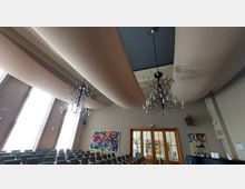 Ein moderner Veranstaltungsraum mit Reihen von gepolsterten Stühlen, großen fensterartigen Wandöffnungen, hängenden Kronleuchtern und wellenförmigen Stoffbahnen an der Decke. An den Wänden sind bunte Gemälde und eine Glastür zu sehen.