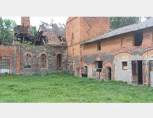 Verfallenes Gebäude aus Backstein und Naturstein mit teilweise eingestürztem Dach, großen Rundbogenfenstern und offenen Türen. Im Vordergrund eine Grasfläche, im Inneren des Gebäudes sind Ziegen sichtbar.