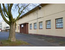 Eine gelbe Backsteinhalle mit großen, vergitterten Fenstern und einem braunen Vordach über dem Eingang, umgeben von einem asphaltierten Hof mit einem großen, kahlen Baum im Vordergrund. Im Hintergrund sind moderne Gebäude sichtbar.