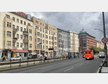 Straßenszene mit mehrstöckigen Wohn- und Geschäftsgebäuden im Berliner Stil, hell verputzt oder in Rotklinkeroptik. Im Vordergrund eine zweispurige Straße, Straßenbahngleise und der Eingang zur U-Bahn-Station "Rosa-Luxemburg-Platz"; ein Krankenwagen fährt auf der Straße.