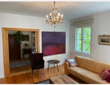 Wohnzimmer mit Holzboden und hellen Wänden, ausgestattet mit einem beigen Ledersofa, einem runden Beistelltisch und einem lila Sessel. Ein großer abstrakter lila- und pinkfarbener Kunstdruck hängt an der Wand; daneben ein Fenster mit Blick ins Grüne und ein Kronleuchter an der Decke. Im Hintergrund ist ein angrenzender Raum mit Bücherregalen und einem Holzschrank sichtbar.