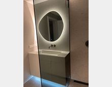 Badezimmer mit modernem Waschbecken, eingelassener Wandarmatur und beleuchtetem, organisch geformtem Spiegel. Die Wände und der Boden sind mit hellen, texturierten Materialien gestaltet.