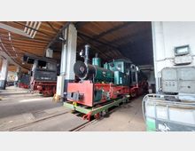 Historische Lokomotive in einer Werkstatthalle mit Holzdach und Betonstützen, umgeben von technischen Geräten und weiteren Zügen im Hintergrund. Die Dampflok ist grün-schwarz mit roten Details und steht auf einem Schienenabschnitt.
