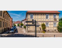 Kopfsteingepflasterte Altstadtstraße mit parkenden Autos und historischen Gebäuden, darunter ein zweigeschossiges Fachwerkhaus mit roten Ziegeldach und Blumenkästen. Im Vordergrund steht ein Wegweiser mit Hinweisen unter anderem zum Bahnhof und zur Touristinformation.