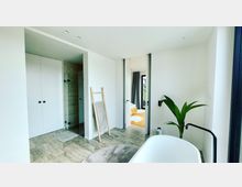 Modernes Badezimmer mit einer freistehenden Badewanne, großen weißen Wänden und hellen Fliesenböden. Im Hintergrund sind eine Dusche mit Glaswand, ein Holzhandtuchhalter sowie der Zugang zu einem angrenzenden Raum mit Holzboden und bodentiefem Fenster erkennbar.