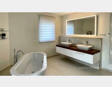 Modernes Badezimmer mit freistehender Badewanne und Armatur im Vordergrund sowie Doppelwaschtisch auf weißem Unterschrank mit Holzplatte. An der Wand hängt ein großer, hinterleuchteter Spiegel; daneben befinden sich ein Fenster mit Rollo und ein hoher Handtuchheizkörper.