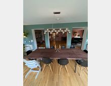 Esszimmer mit einem großen holzfarbenen Tisch und acht Stühlen mit hellen Holzbeinen. An der Decke hängt eine moderne Hängeleuchte mit kugelförmigen Lampen, und im Hintergrund sind angrenzende Zimmer mit unterschiedlich gefärbten Wänden sichtbar.