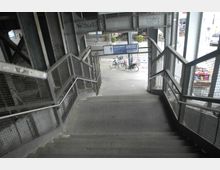 Eine Treppe unter einer Bahnüberführung mit Metallgeländern und Gitterwänden, die zu einem Gehweg führt. Im Hintergrund sind Fahrräder, Hinweisschilder und Straßenszenen erkennbar.