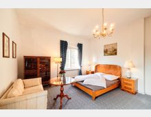 Helles Schlafzimmer mit Doppelbett aus Holz, Nachttischen mit Lampen und Kronleuchter an der Decke. Vor einem Fenster mit dunklen Vorhängen stehen ein runder Glastisch, ein Sofa und ein Holzvitrinenschrank an der Wand.