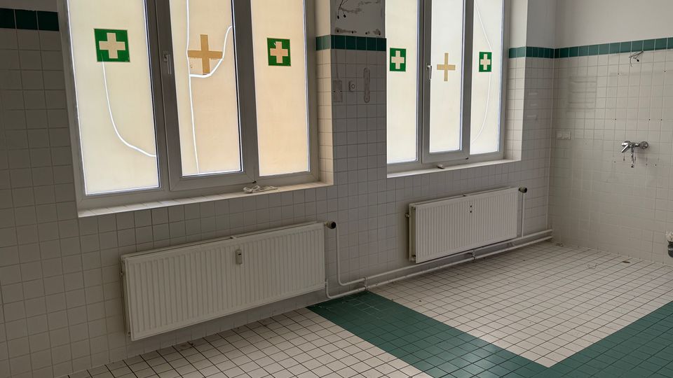 Gefliester Innenraum mit weiß-grauen Wandfliesen und grüner Bordüre, zwei großen Milchglasfenstern mit Erste-Hilfe-Kreuzsymbolen sowie Heizkörpern darunter. Auf dem Boden liegen weiße Fliesen mit einem grünen Streifen; an der rechten Wand sind Wasseranschlüsse sichtbar.