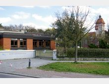 Ein flaches Schulgebäude aus rotem Klinker mit großen Fenstern, umgeben von Bäumen und begrenzt durch einen Zaun. Im Hintergrund ist ein historisches Gebäude mit einem Turm und rotem Ziegeldach sichtbar.