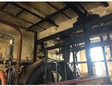 Industriehalle mit alter Maschinenanlage; sichtbare Elemente umfassen Rohrleitungen, Zahnräder, einen Generator und Stahlträger in einer abgenutzten Umgebung mit sichtbarer Patina an den Wänden und Decken. Licht dringt durch ein Fenster im Hintergrund, das den Raum schwach beleuchtet.