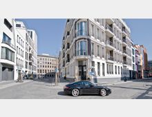 Straßenecke in einem städtischen Gebiet mit modernen Wohn- und Bürogebäuden, darunter ein Eckgebäude mit Balkonen und großen Fenstern. Im Vordergrund ist ein schwarzer Sportwagen auf der Straße zu sehen, bei klarem Himmel und ruhigem Verkehr.