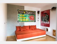 Ein modern eingerichtetes Zimmer mit einer orangen Tagesliege, Wandpostern (eins mit der Aufschrift „Trip to Jamaica“) und einem Holzboden. Eine Betonwand und ein Regal auf der rechten Seite tragen zur minimalistischen Dekoration bei.