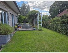 Eine Terrasse mit Sitzmöbeln aus Rattan neben einem Haus mit Klinkerfassade, umgeben von einem gepflegten Garten mit Grasfläche, Hecken und Bäumen. Im Hintergrund ist ein Holzgerüst mit einem Hängesessel angebracht, und der Himmel ist teilweise bewölkt.