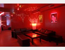 Ein Lounge-Bereich mit roten Wänden, schwarzem Ledersofa, Holztisch und rotem indirektem Licht. Die Wände sind mit leuchtenden Dekorationen und Postern geschmückt, und der Raum hat einen Holzboden.