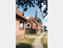 Backsteinkirche mit rotem Ziegeldach und hohem, spitzem Kirchturm, umgeben von einer Wiese. Ein gepflasterter Weg führt entlang der Kirche, im Hintergrund sind Bäume und weitere Gebäude zu sehen.
