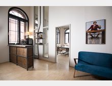 Heller Innenraum eines modernen Büros oder Wohnraums mit großen, schwarzen Rundbogenfenstern. Zu sehen sind eine Holzzähler mit Kaffeemaschine, ein blauer Sessel, ein Poster an der Wand und ein Durchgang zu einem weiteren Raum mit Tisch und Stühlen.