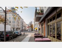 Straßenszene in einer Wohngegend mit mehrstöckigen Altbauten, Bäumen am Straßenrand und parkenden Autos. Im Vordergrund rechts ein Restaurant mit Terrassentischen und rot-weiß karierten Tischdecken; im Hintergrund ein Stoppschild und eine Querstraße.