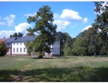 Ein weißes Gebäude mit großen, symmetrischen Fenstern und einem dunklen Schieferdach steht in einer grünen Parklandschaft. Im Vordergrund befindet sich eine Wiese, umgeben von vereinzelten Bäumen, und der Himmel ist leicht bewölkt.