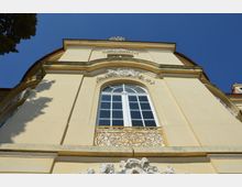 Nahaufnahme einer gelb gestrichenen Barockfassade mit einem großen, bogenförmigen Fenster und dekorativen Verzierungen aus weißem Stuck. Unter dem Fenster befindet sich ein schmiedeeisernes Geländer in Goldfarbe, und der Himmel ist klar und blau.