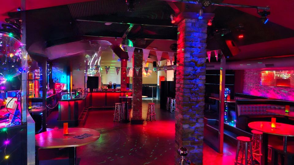 Innenraum einer Bar oder eines Clubs mit bunten LED- und Spotlichtern, Bar-Theke im Hintergrund und mehreren hohen Tischen mit Barhockern. Sichtbar sind eine Diskokugel, Steinpfeiler, eine abgehängte Decke sowie Sitznischen entlang der Wand.