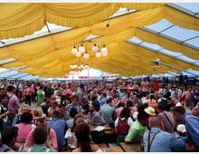 Ein gefülltes Festzelt mit gelb dekoriertem Stoffhimmel, einer großen Menschenmenge in Trachtenkleidung, langen Holzbänken und Tischen sowie dekorativen Hängelampen. Im Hintergrund befinden sich Verkaufsstände und eine Bühne mit Musikern.